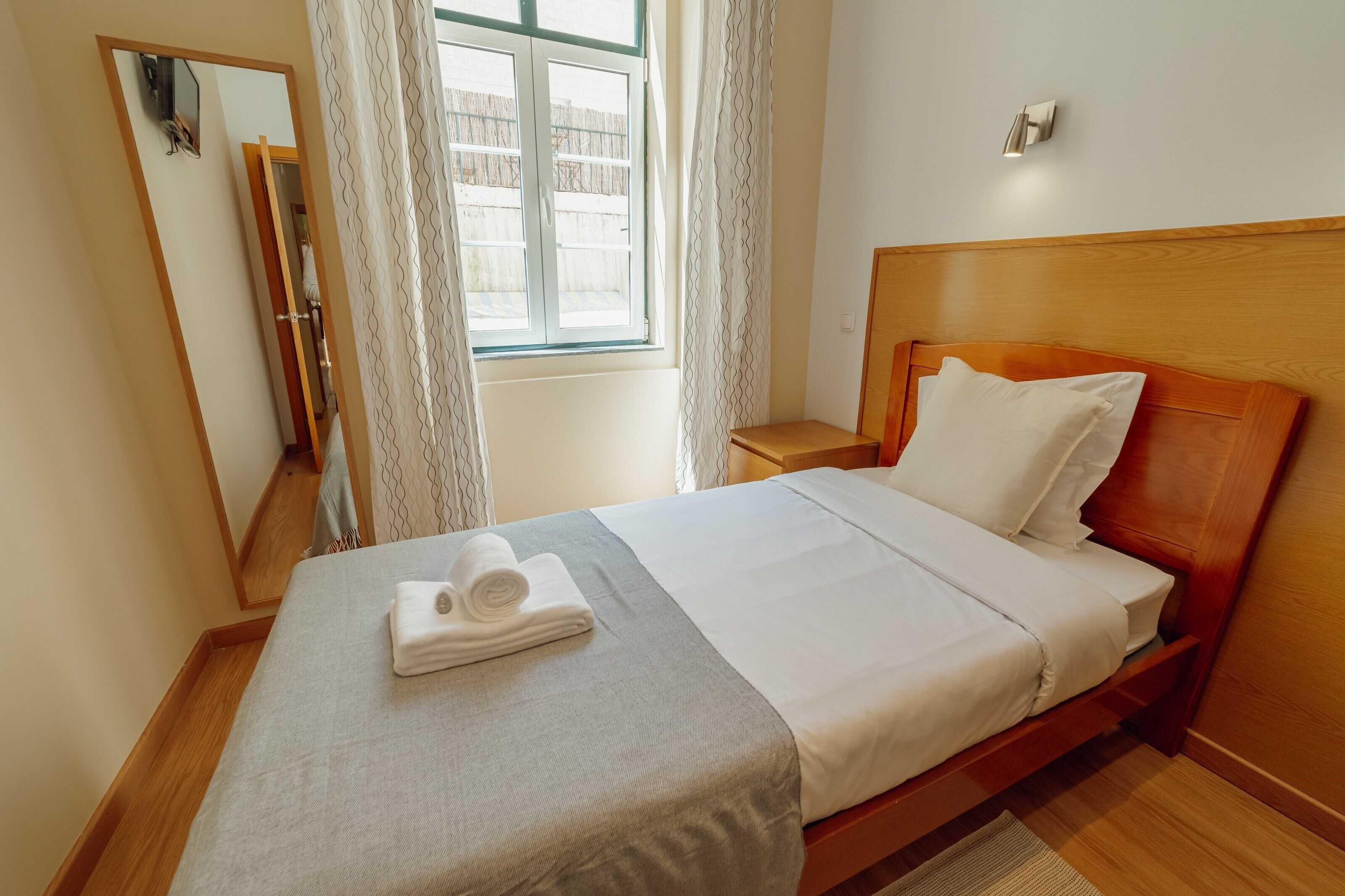 A neatly made bed with a folded towel, a bedside table, a mirror, and a window with curtains.