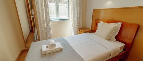 A neatly made bed with a folded towel, a bedside table, a mirror, and a window with curtains.