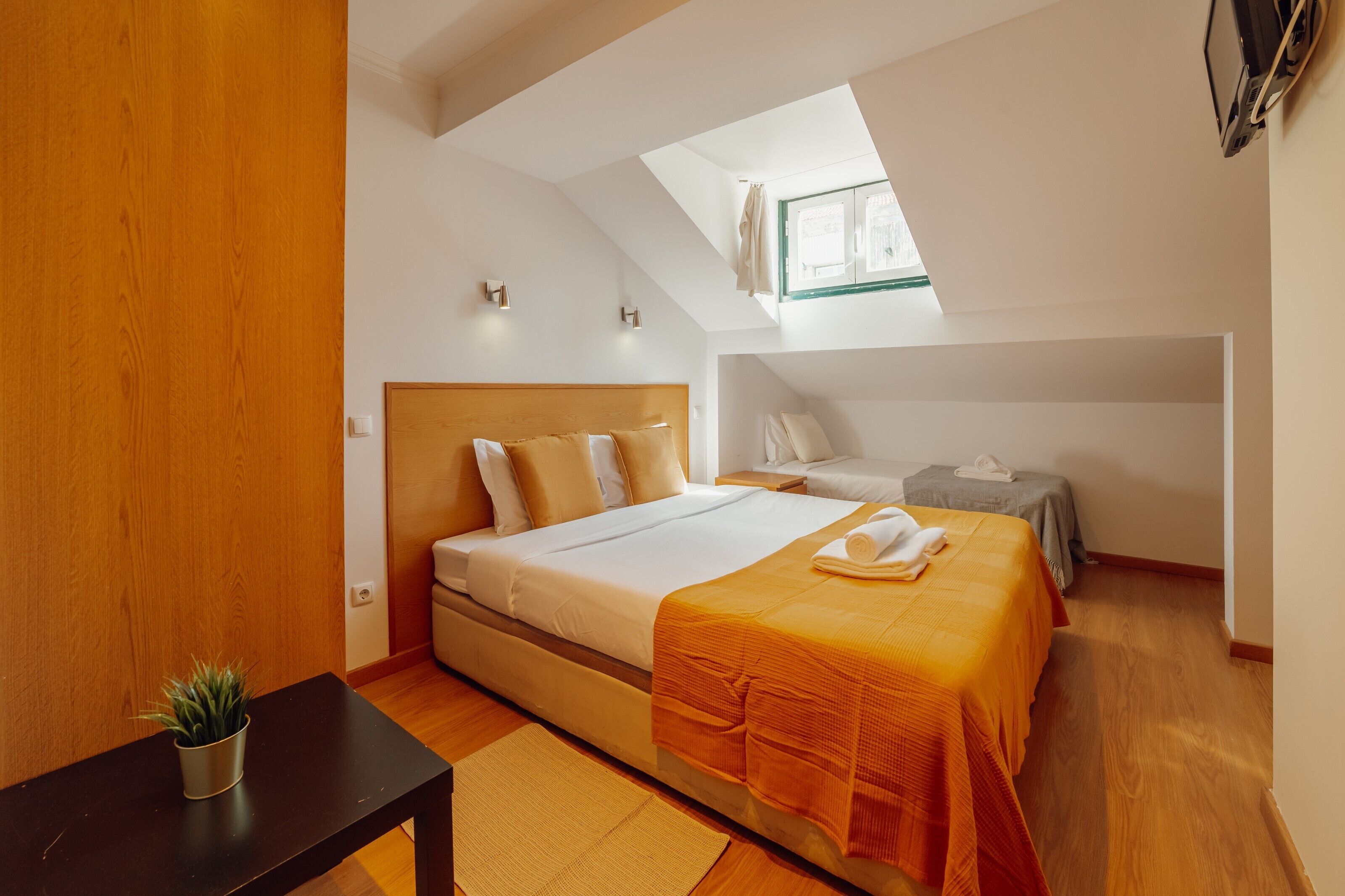 Une chambre à coucher avec un lit, une petite table sur laquelle est posée une plante, une fenêtre et une lucarne.