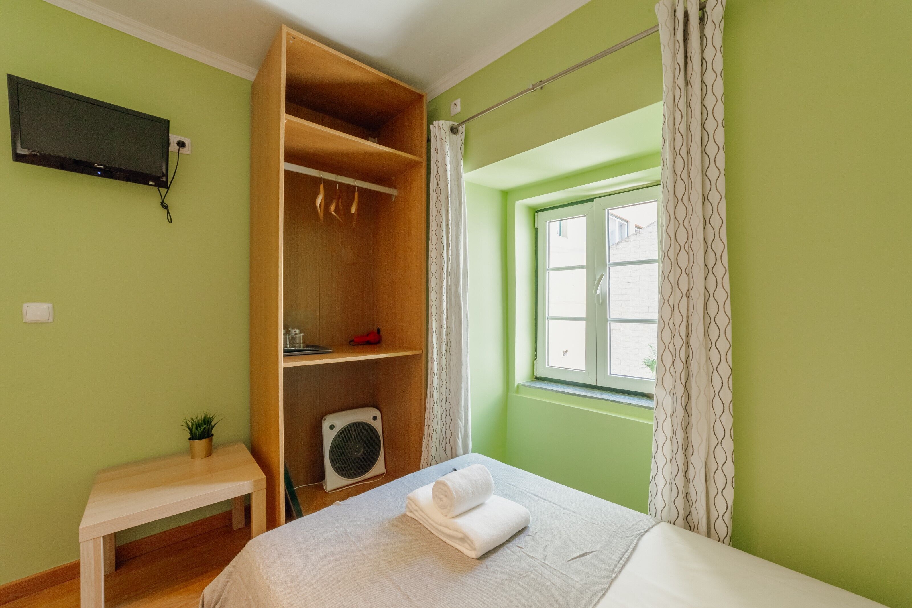 Ein Zimmer mit einem Bett, einem Holzschrank, einem kleinen Tisch mit Pflanze, einem Ventilator und einem an der Wand befestigten Fernseher.