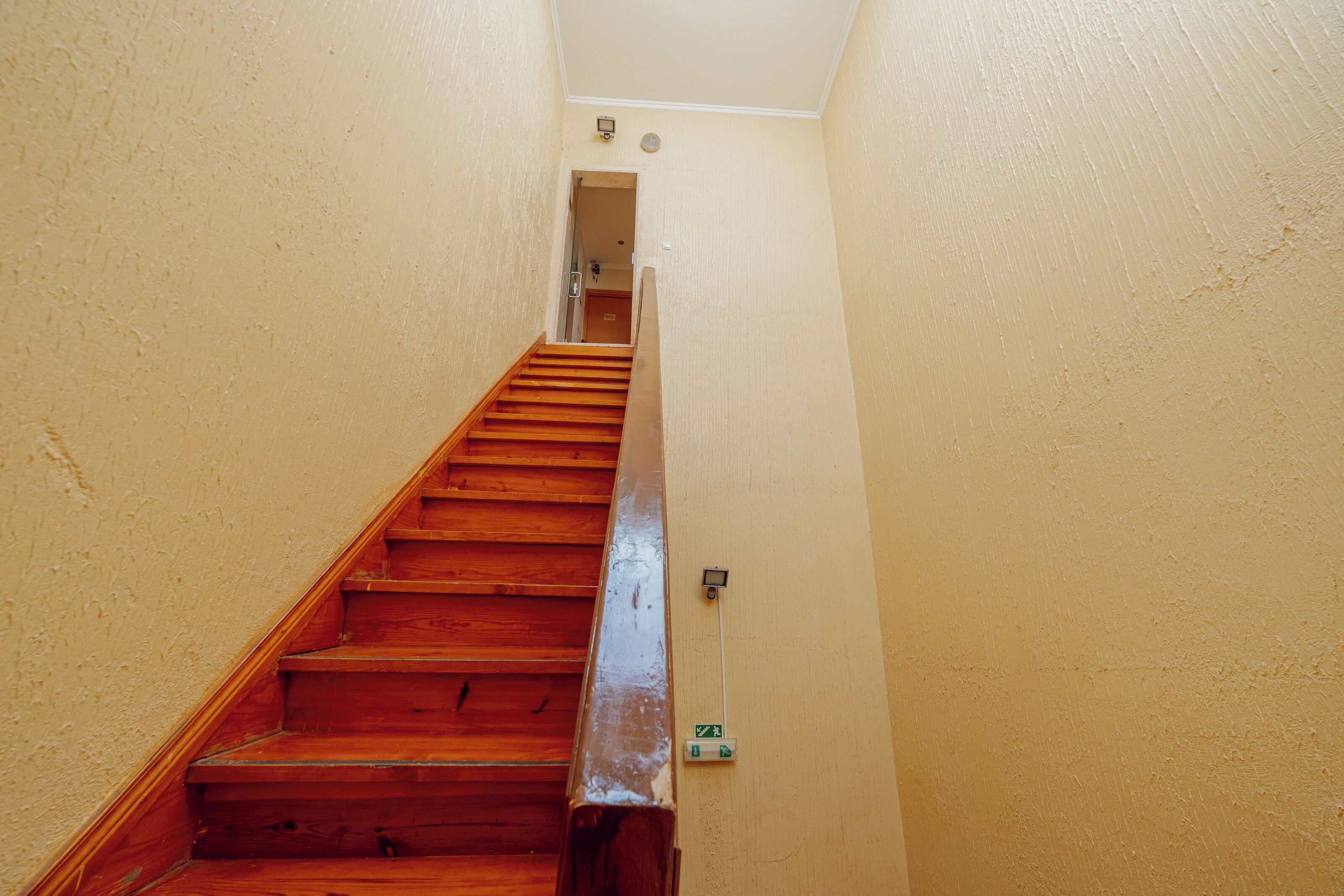 Un escalier étroit avec des marches en bois menant à un palier, des murs de couleur beige et un interrupteur sur le mur.