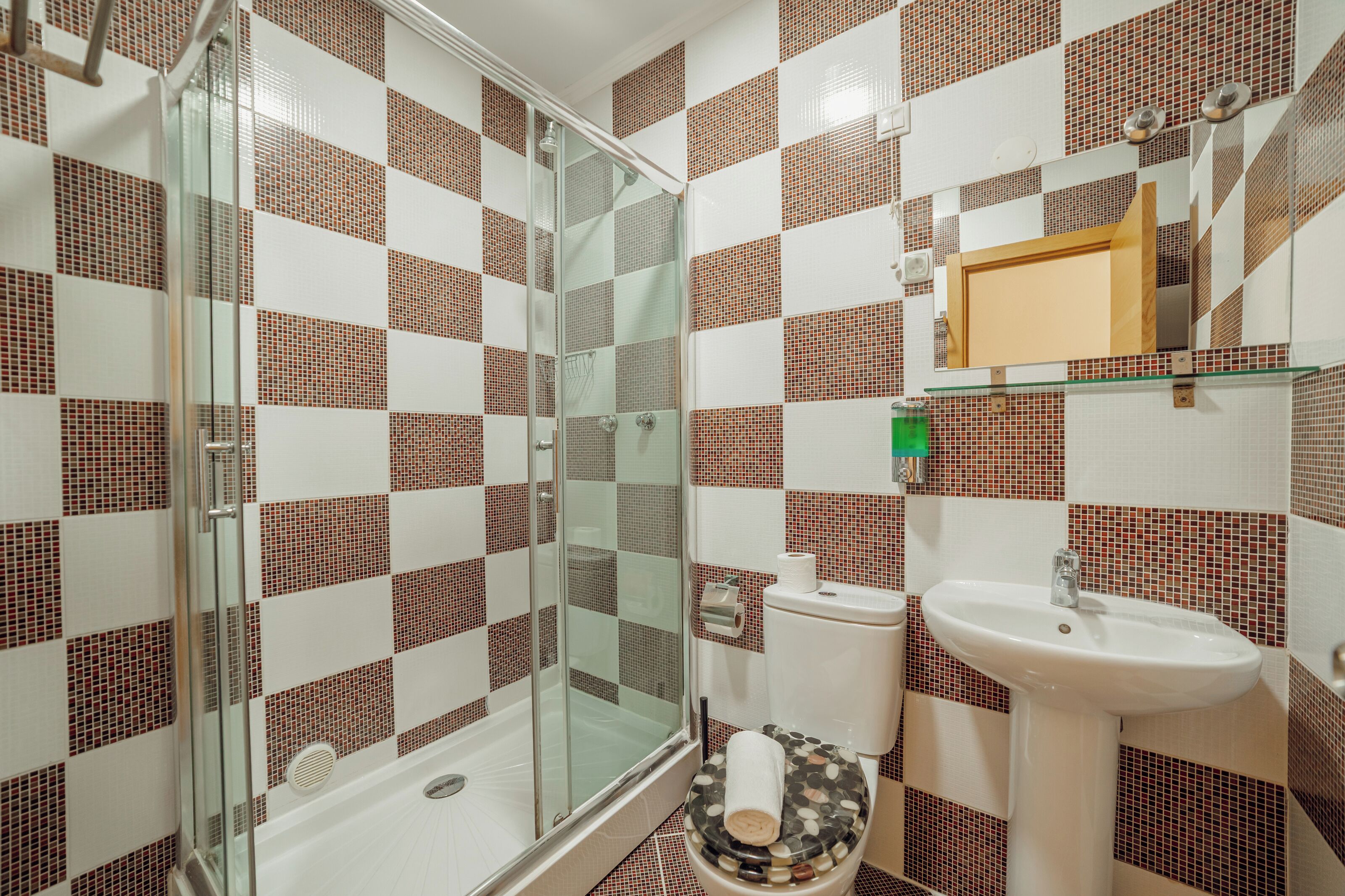 Ein Badezimmer mit Duschkabine aus Glas, einem weißen Waschbecken und einer Toilette mit Mosaikfliesen-Verkleidung.