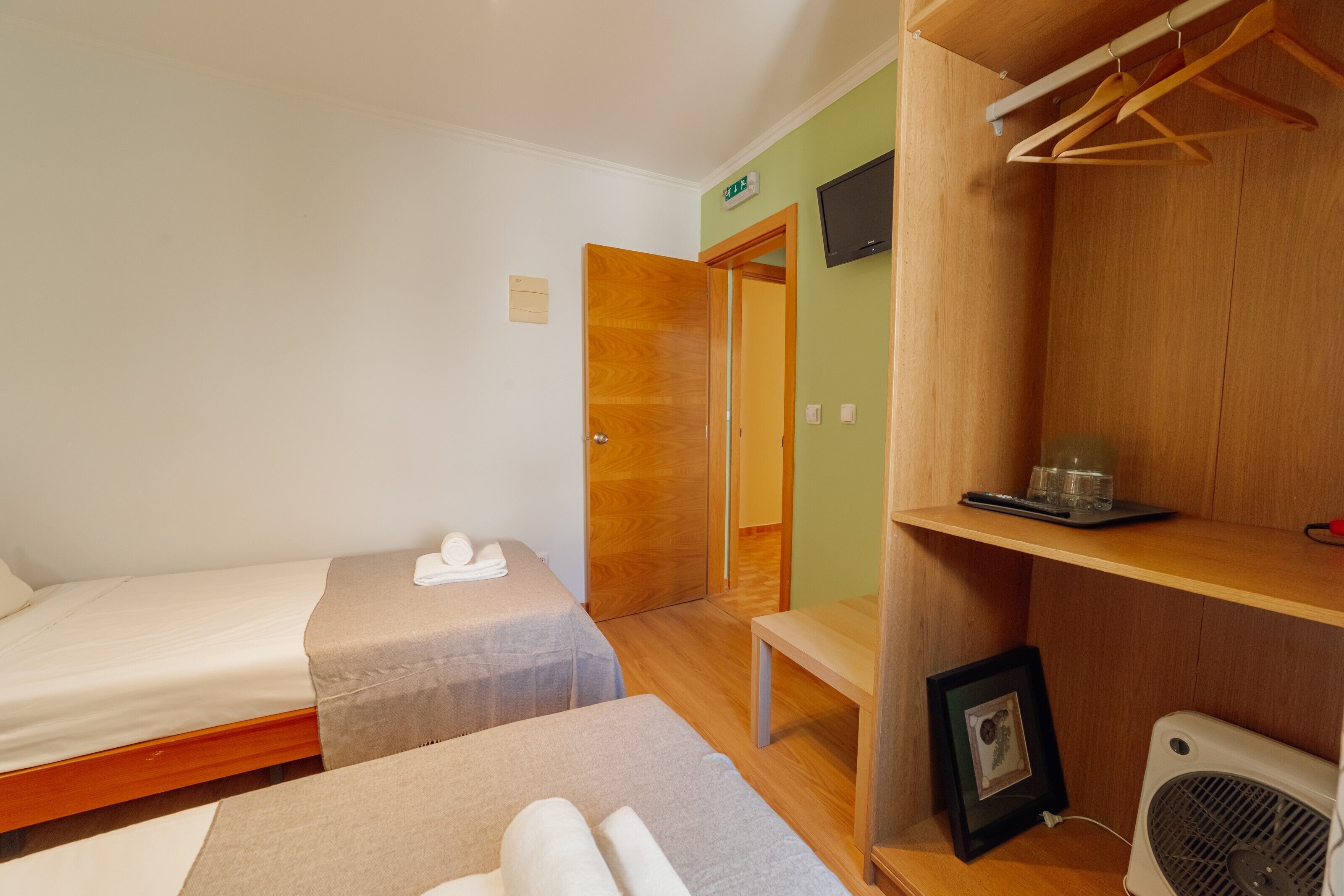 A room with two beds, a wooden wardrobe, a small table, a framed picture, and a fan.