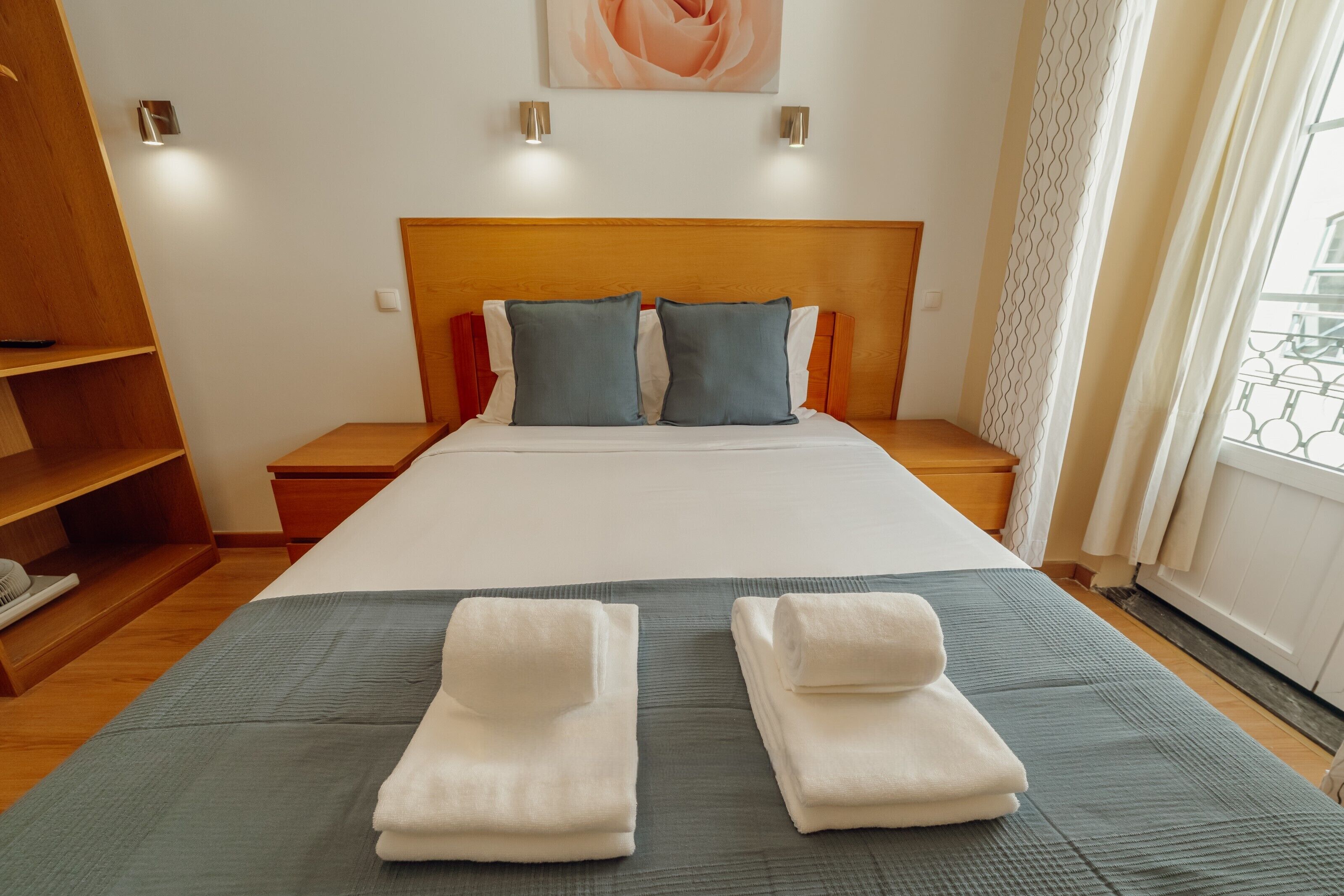 A neatly made bed with two pillows, a wooden headboard, a nightstand, a shelf with a toothbrush, and a window with curtains.