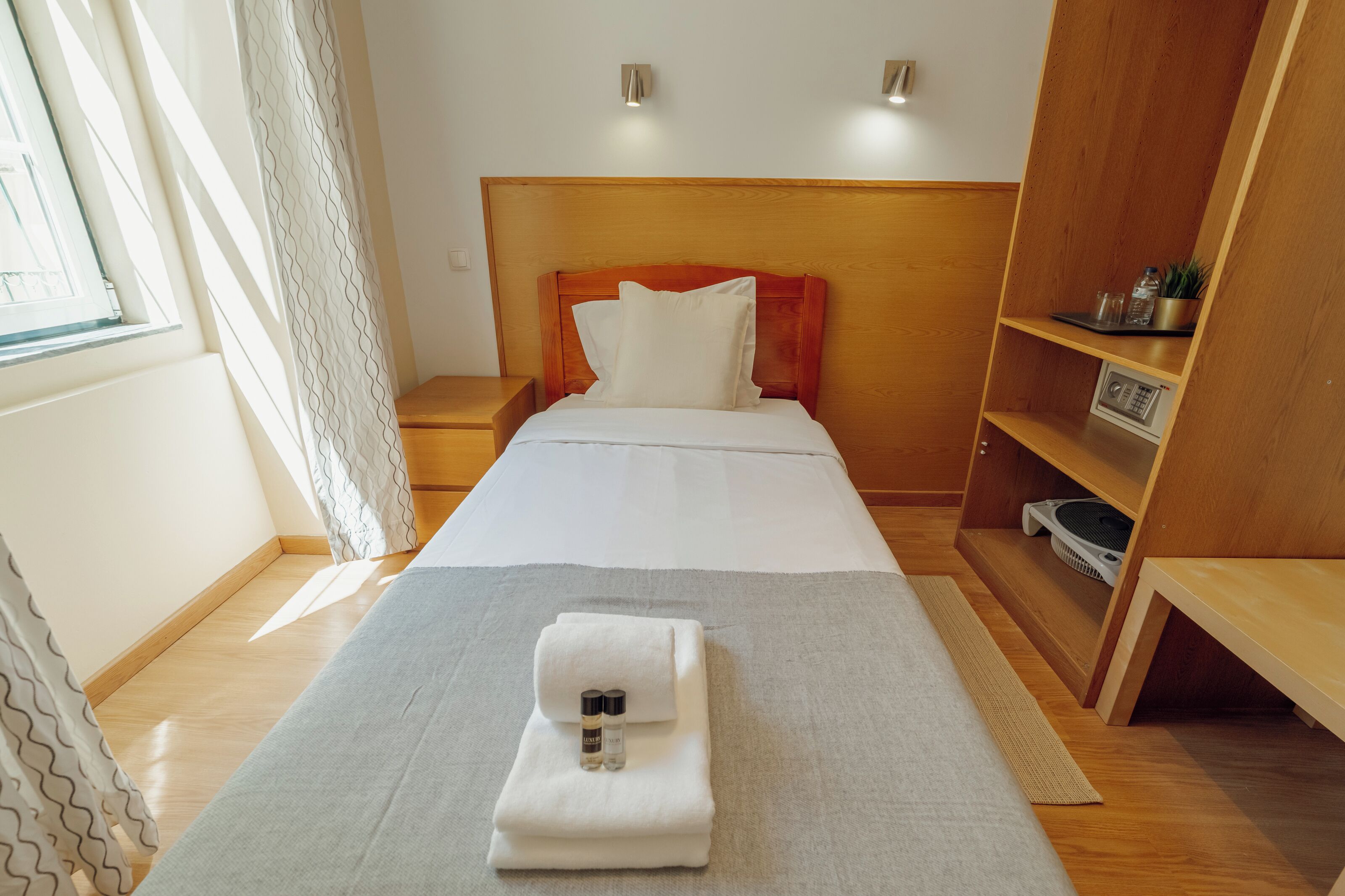 A hotel room with a bed, a bedside table, a nightstand, a wooden wardrobe, and a window with curtains.