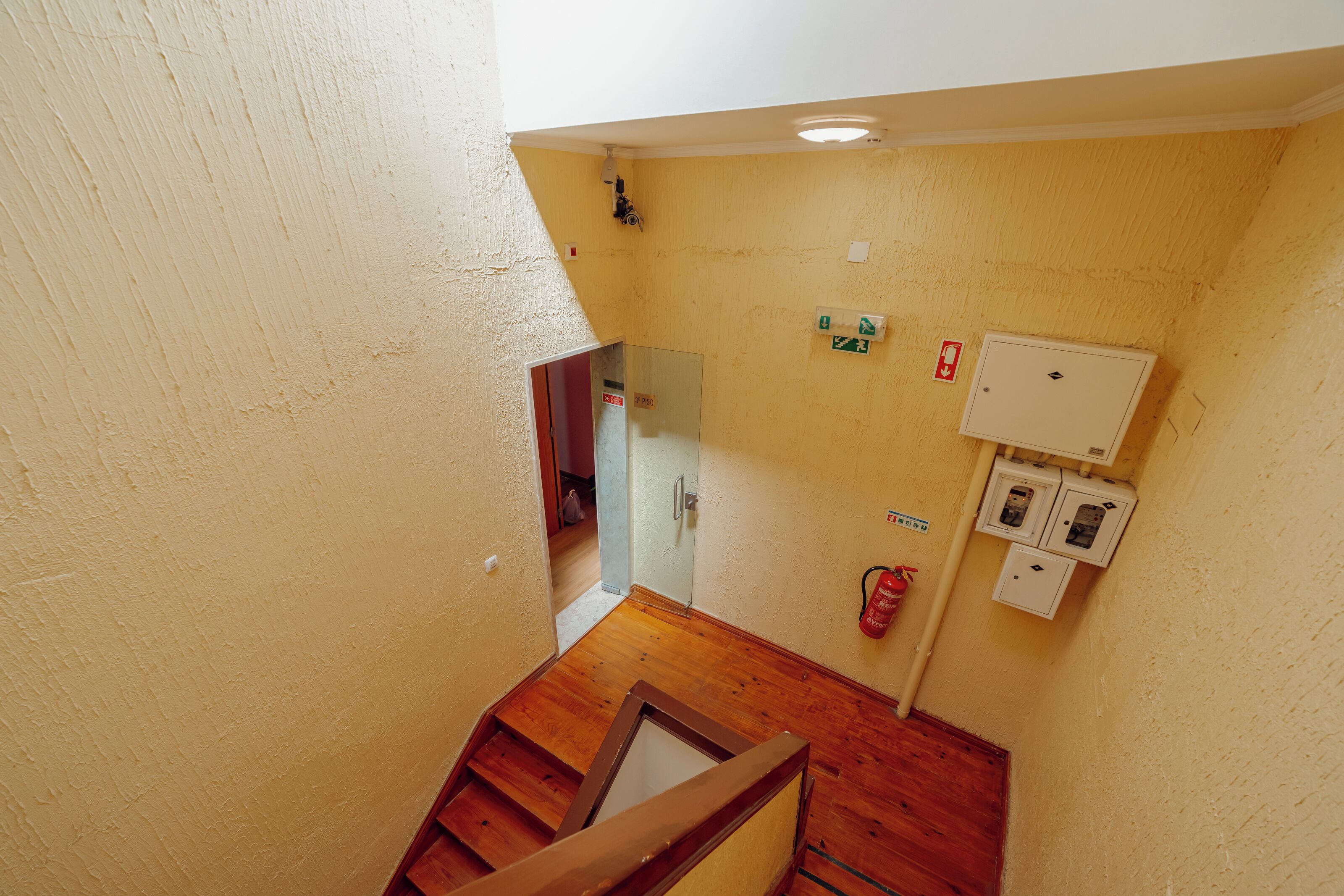 A stairwell with a wooden handrail, a fire extinguisher, and electrical boxes on the wall.
