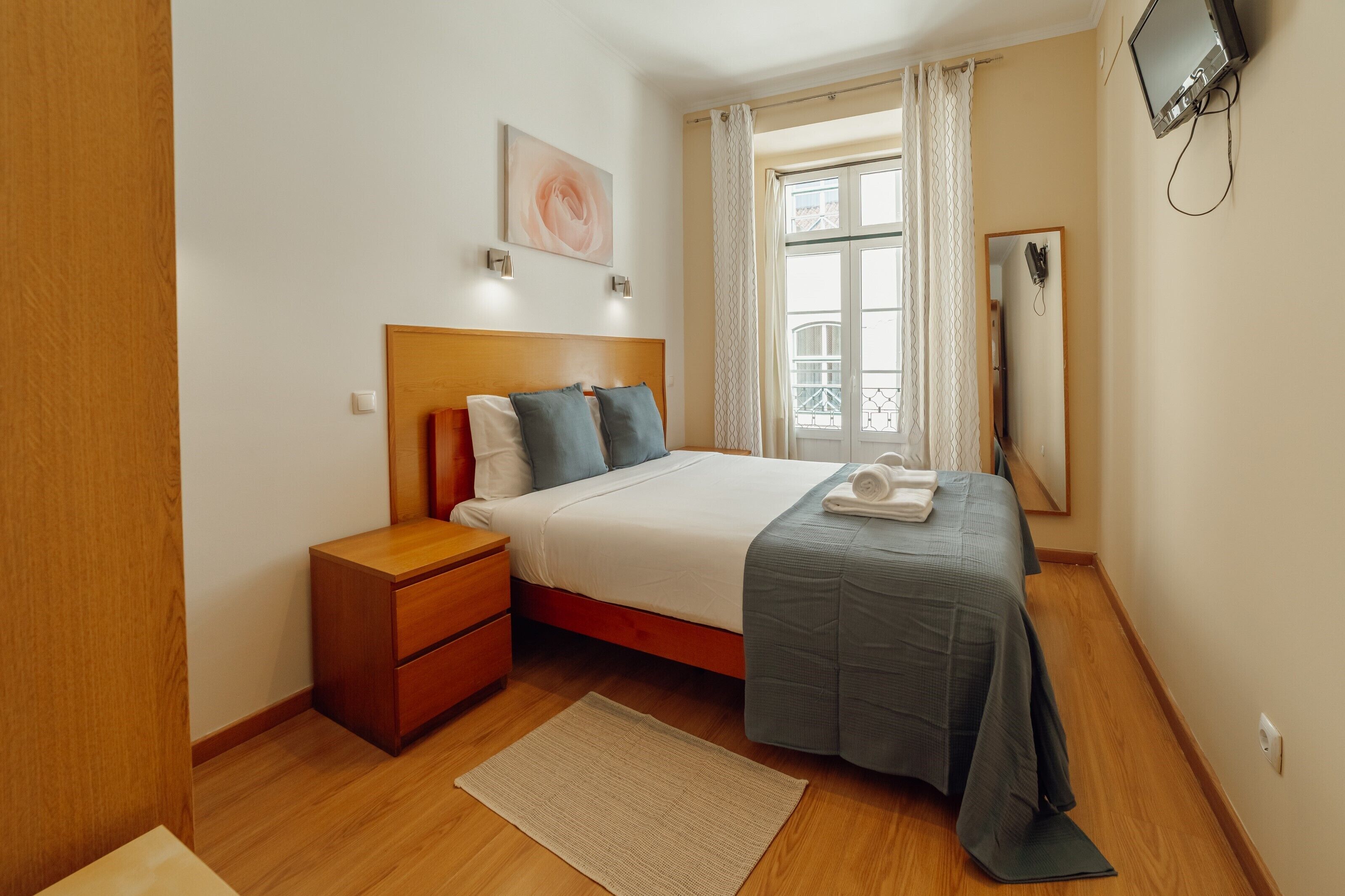 A bedroom with a bed, nightstand, bedside table, window with curtains, and a wall-mounted TV.