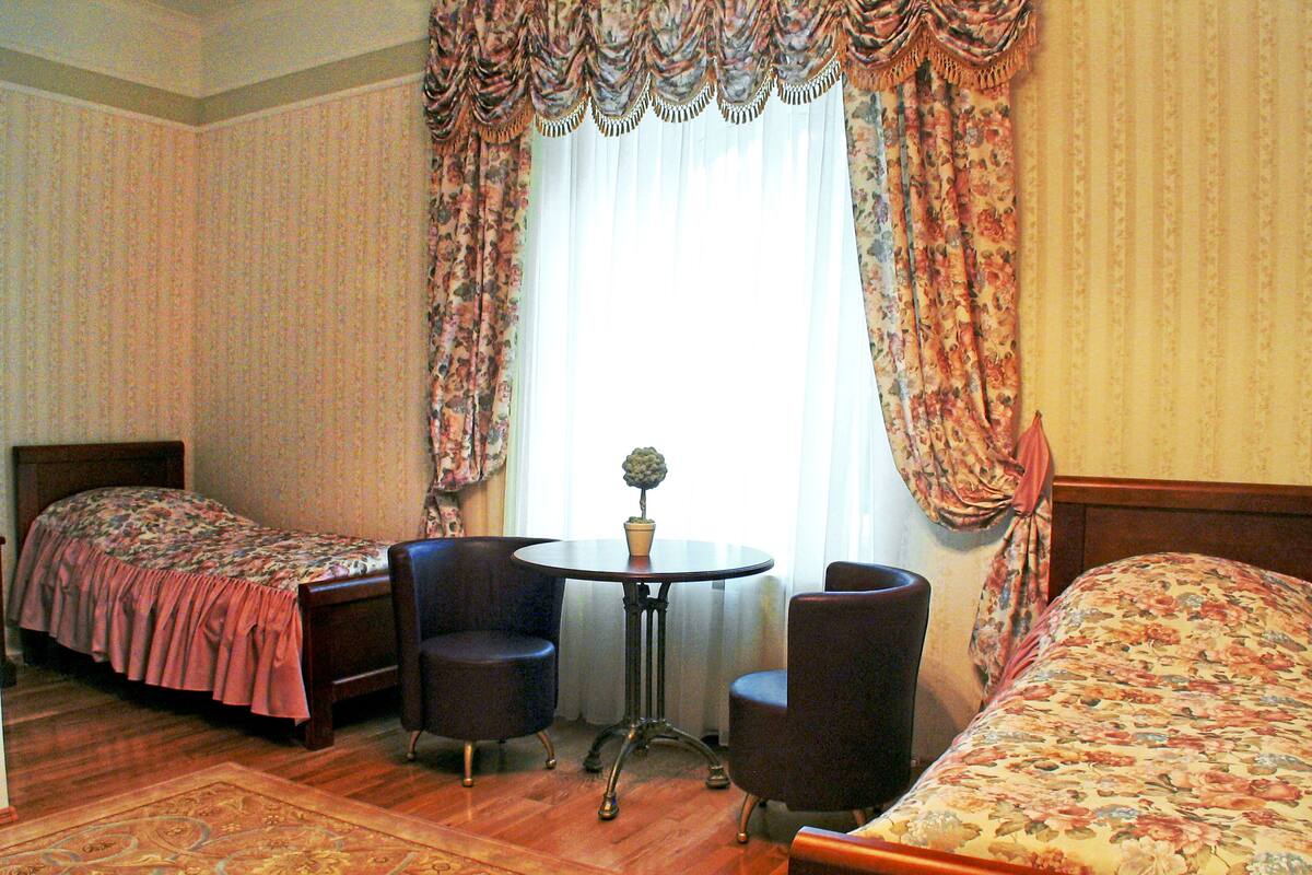 A hotel room with two single beds, a small round table with chairs, a potted plant, and a window with curtains.