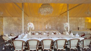 Un salón de banquetes con una larga mesa dispuesta para un evento formal, rodeado de sillas de mimbre.