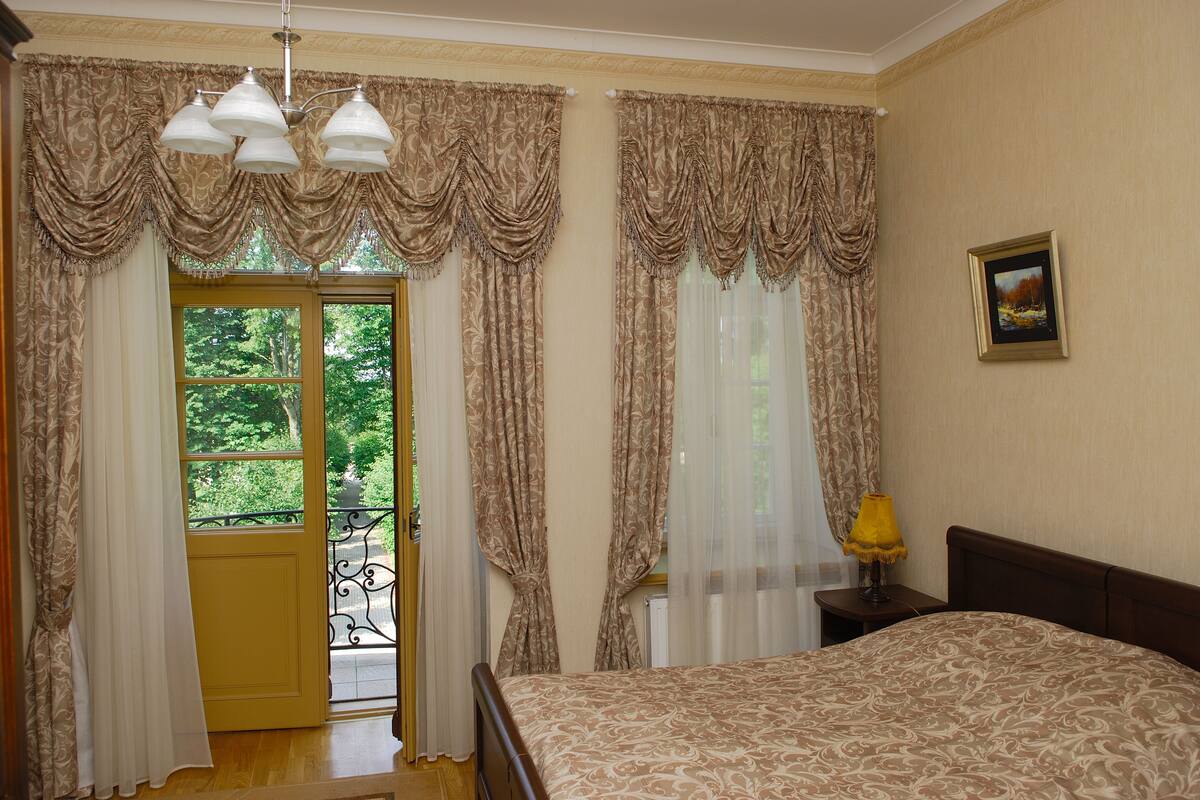 A bedroom with a bed, curtains, a window with a view, a nightstand with a lamp, and a framed picture on the wall.