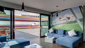 A modern lounge area with a large window, blue sofas, and a view of a colorful building outside. - MEININGER Hotel Berlin Airport (Berlin)