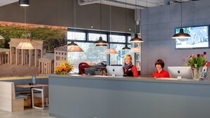 A modern reception area with a large window, a desk with computers, and a seating area with a view of a cityscape mural. - MEININGER Hotel Berlin Airport (Berlin)