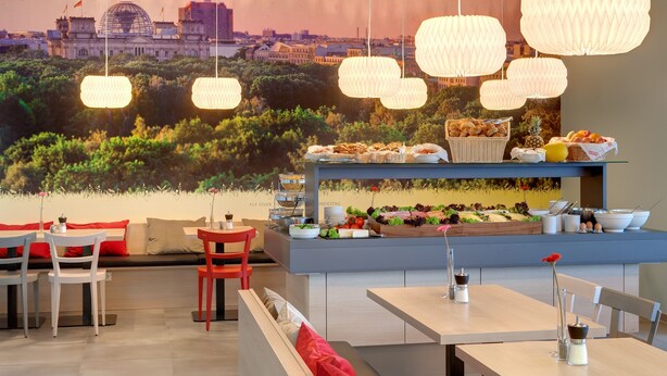 A restaurant with a cityscape mural, hanging paper lanterns, and a buffet with various dishes. - MEININGER Hotel Berlin Airport (Berlin)