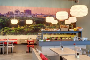 A restaurant with a cityscape mural, hanging paper lanterns, and a buffet with various dishes. - MEININGER Hotel Berlin Airport (Berlin)