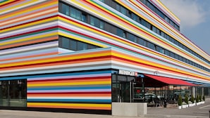 A modern hotel with a colorful striped facade and a covered entrance area with seating. - MEININGER Hotel Berlin Airport (Berlin)