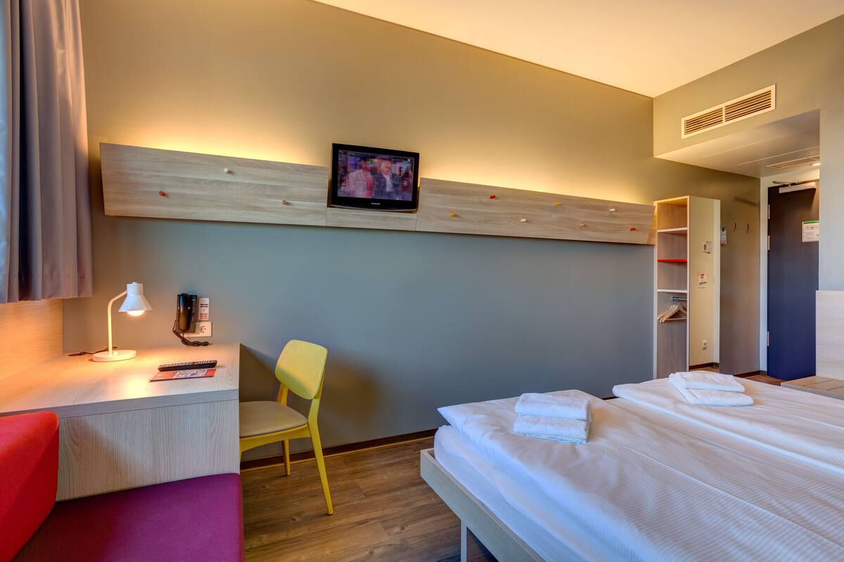 A hotel room with a bed, a desk with a lamp, a chair, a TV mounted on the wall, and a wardrobe.