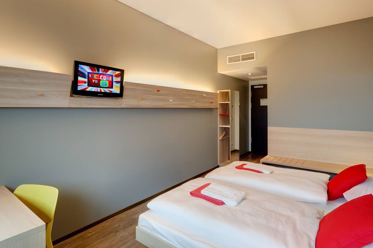A hotel room with a bed, a TV mounted on the wall, a wooden headboard, and a small desk with a chair.