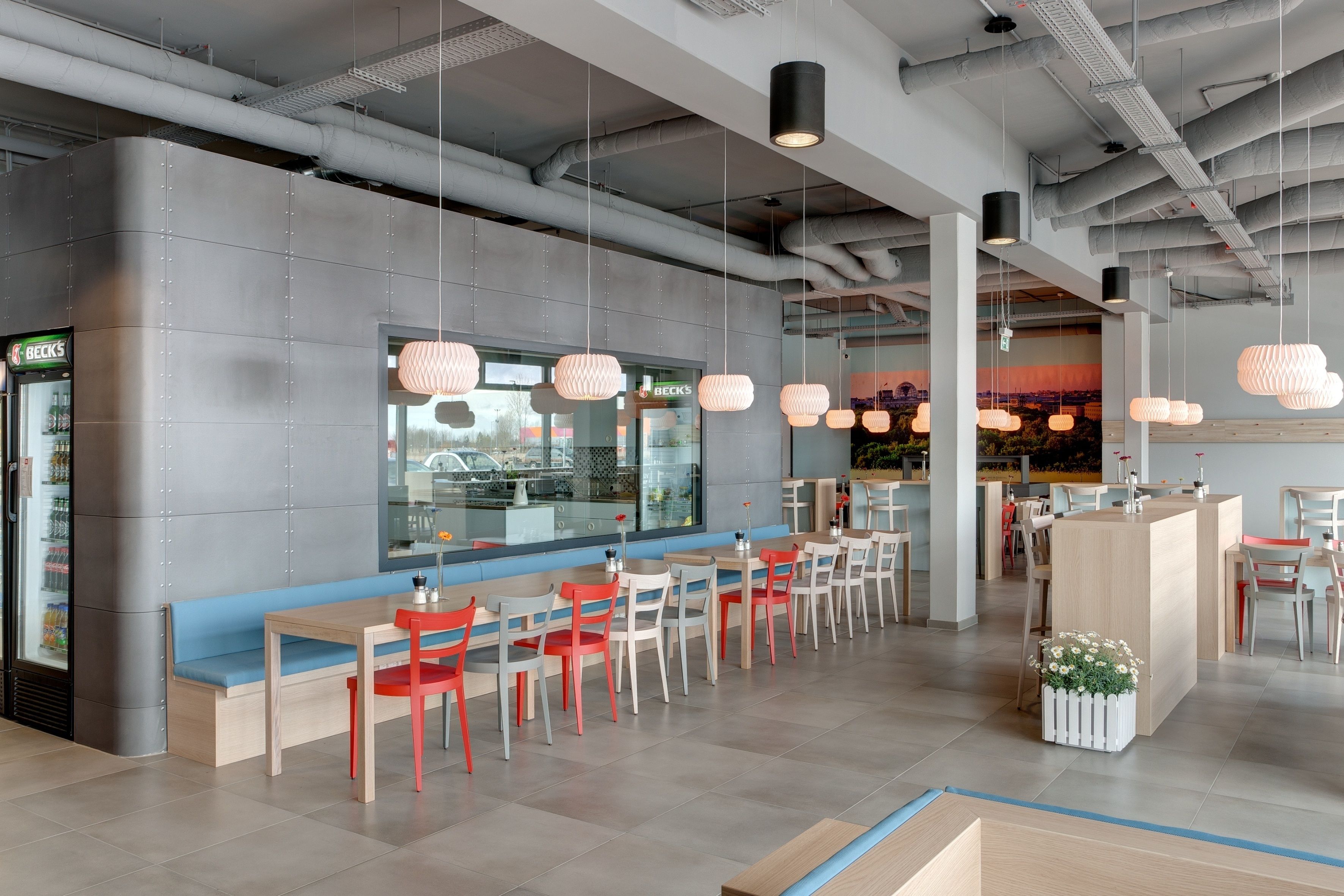 Un restaurante moderno con una larga barra, sillas rojas y blancas, lámparas colgantes y vistas a la ciudad a través de amplias ventanas.