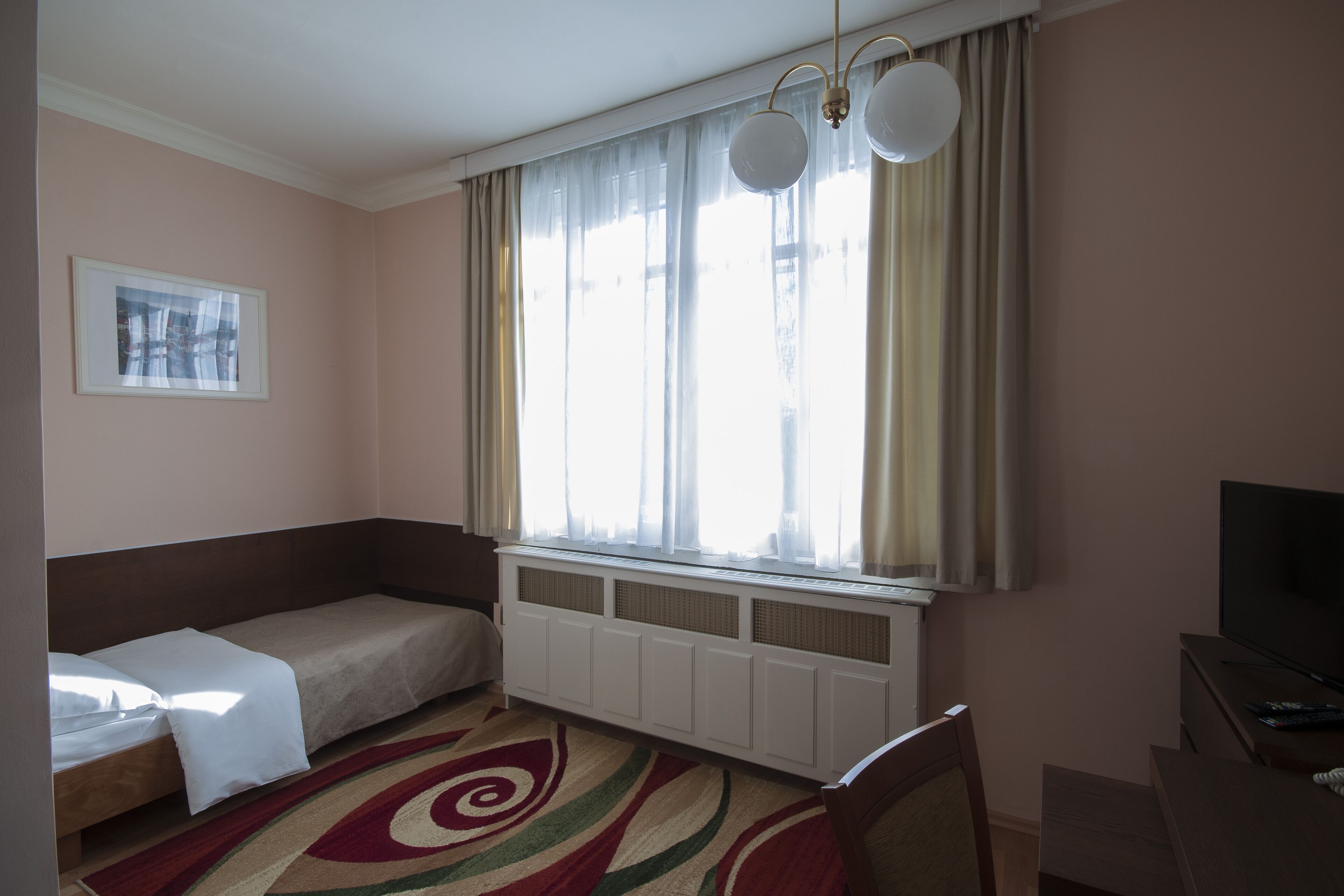 Una habitación pequeña con una cama individual, un radiador, un televisor y una ventana con cortinas.