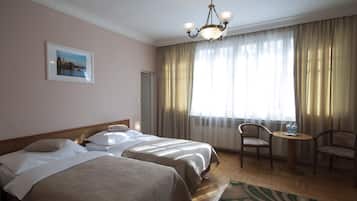 A hotel room with two beds, a chandelier, a painting on the wall, and a view of the outdoors through large curtains.