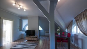 A spacious living area with a wooden floor, a flat-screen TV, a dining table with red chairs, and a large window with curtains.