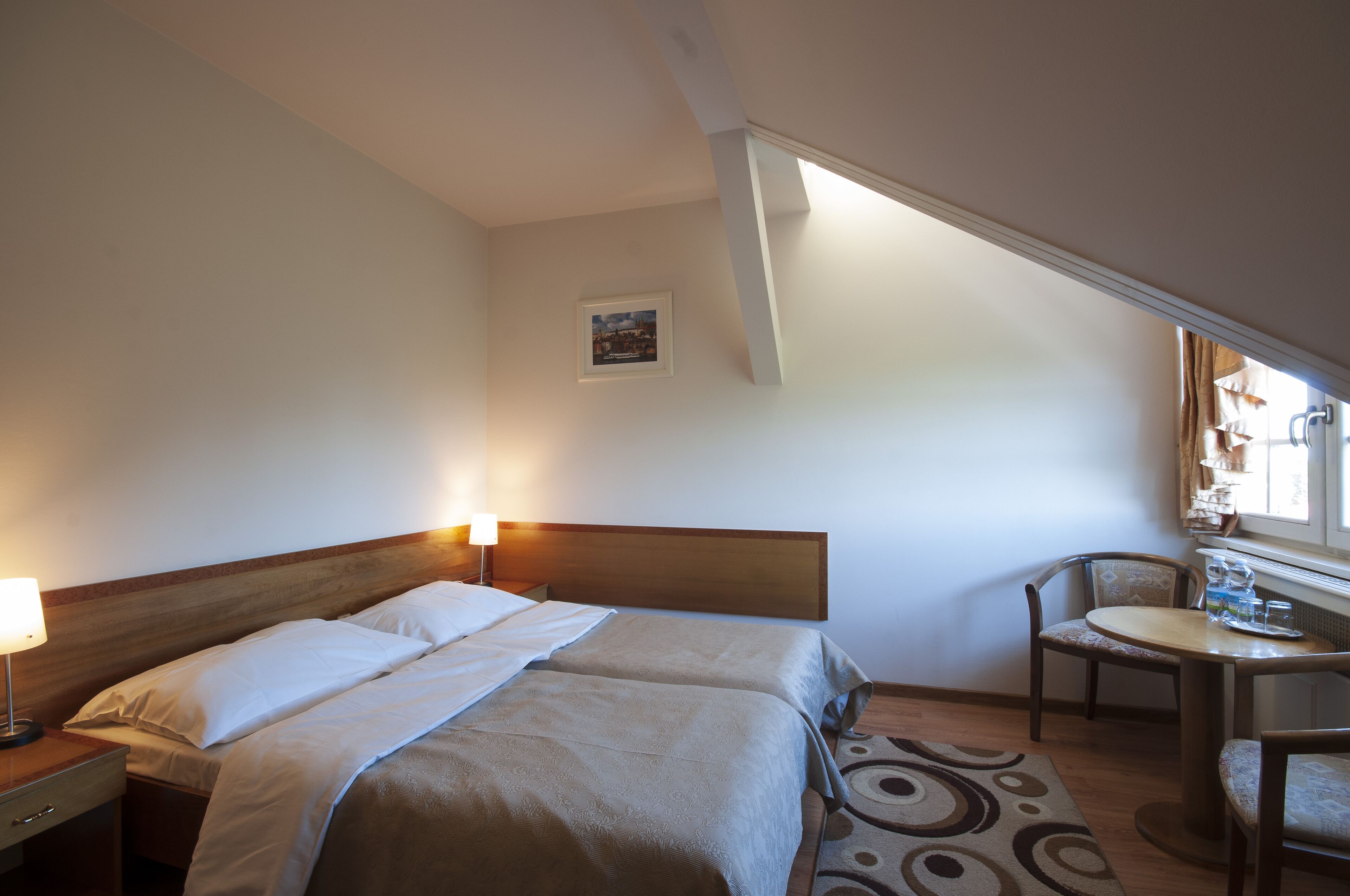Habitación de hotel con cama doble, mesita de noche con lámpara, una pequeña mesa redonda con una silla y ventana con cortinas.