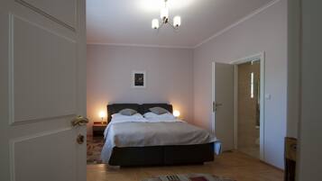 A bedroom with a bed, bedside lamps, a chandelier, and a door leading to another room.