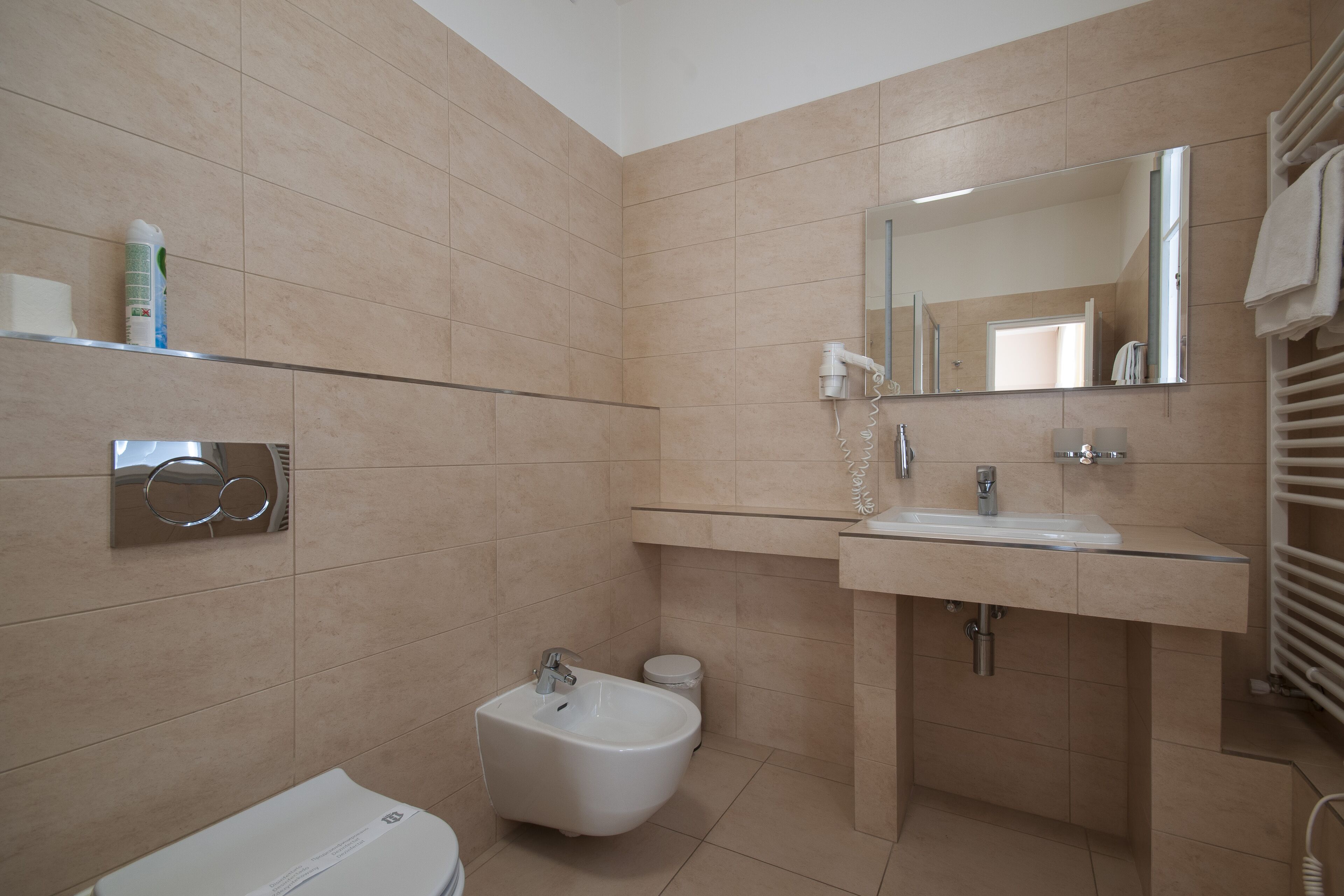 A modern bathroom with beige tiles, a bidet, a sink, a mirror, and a towel rack.