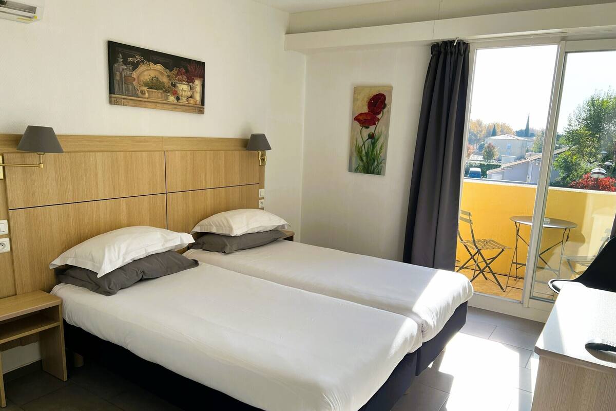 Une chambre d’hôtel avec un lit double, une tête de lit en bois, une petite table de chevet, une lampe, un tableau au mur et une vue sur le balcon extérieur.