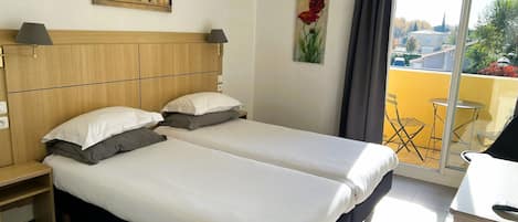 A hotel room with a double bed, a wooden headboard, a small bedside table, a lamp, a painting on the wall, and a view of the outside balcony.