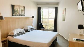 Habitación de hotel con una cama grande, un escritorio con computadora, una silla y una ventana con cortinas.
