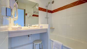Een badkamer met een witte wastafel, spiegel en douche. Er liggen handdoeken op de wastafel en een kruk voor het toilet.