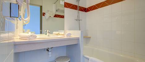Un baño con lavamanos, espejo y ducha blancos. Hay toallas sobre el lavamanos y un taburete frente al inodoro.