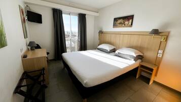 Una habitación de hotel con cama, escritorio, silla, televisor, un cuadro y vistas de la ciudad.