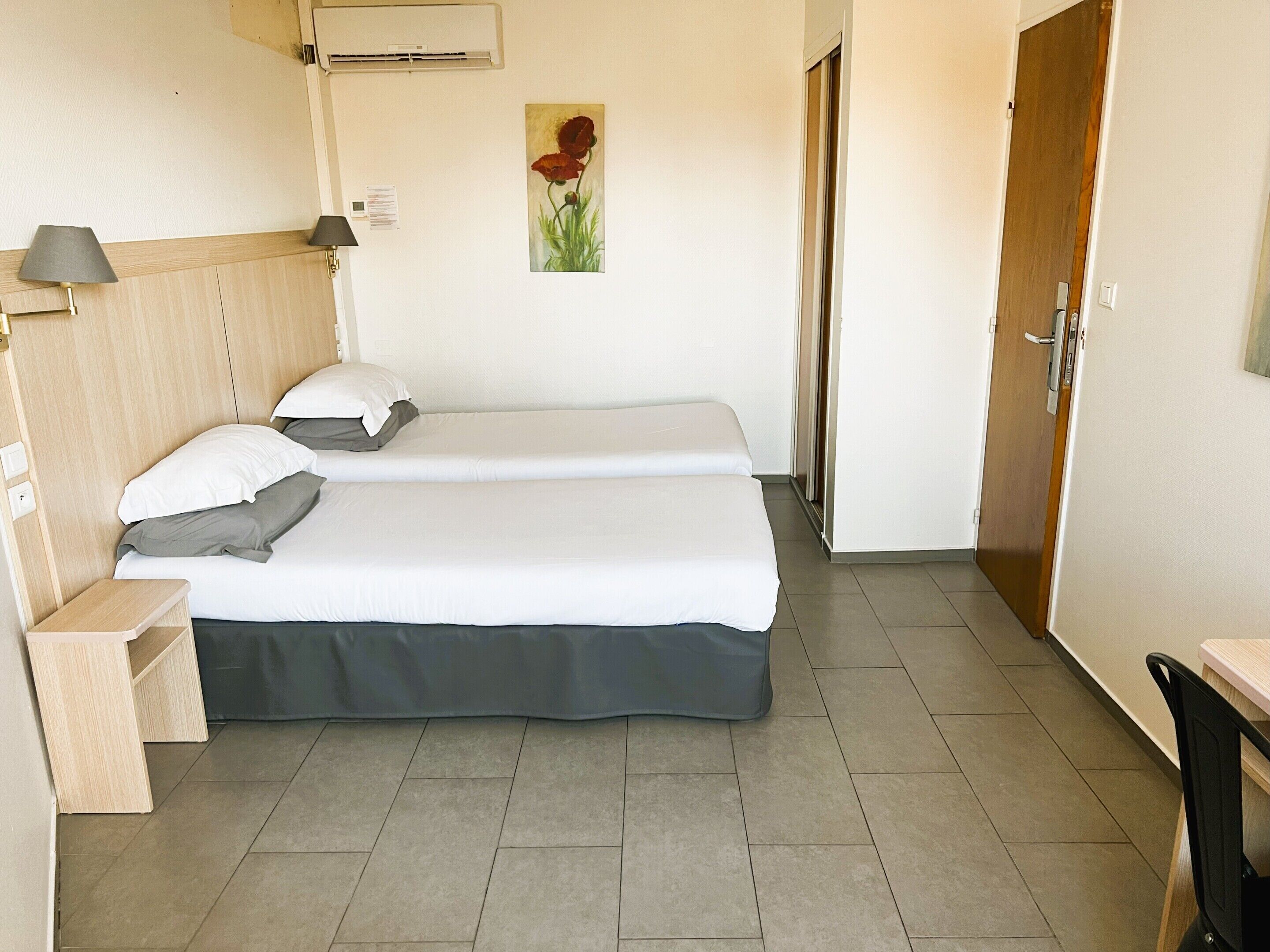 A hotel room with two beds, a wooden headboard, a small bedside table, a lamp, a painting, and a door.