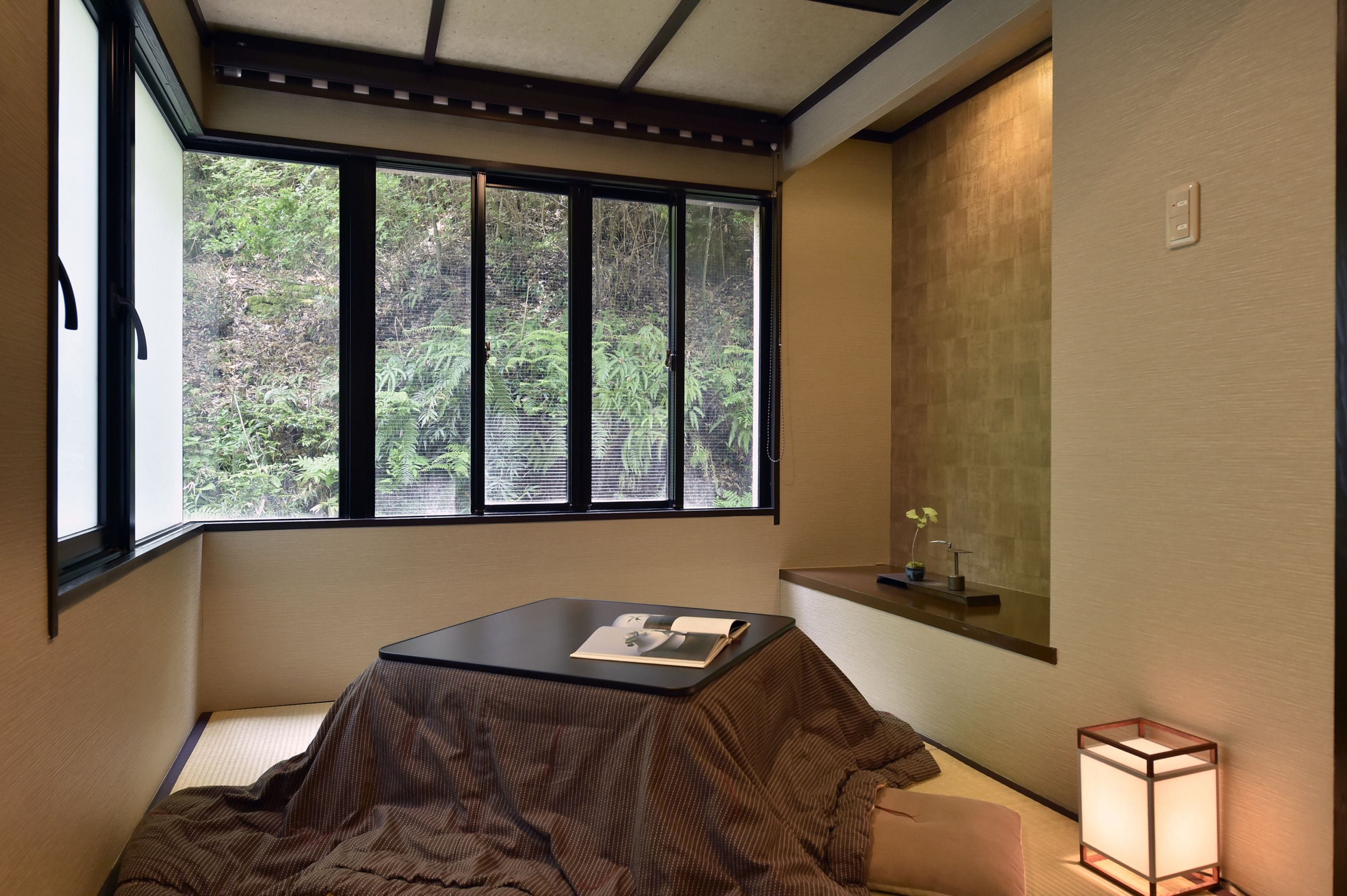 A room with a tatami mat seating area, a table with a book, a window with a view of greenery, and a bedside table with a lamp.