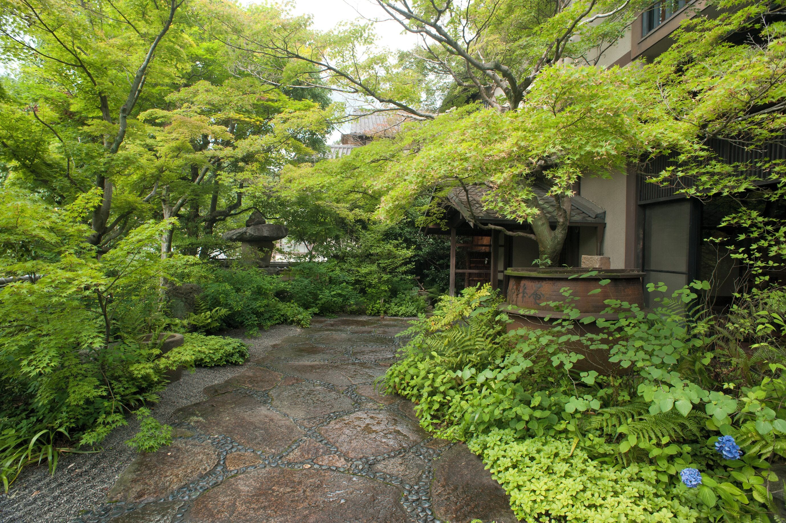 A traditional Japanese garden with a stone pathway, lush greenery, and a wooden structure.