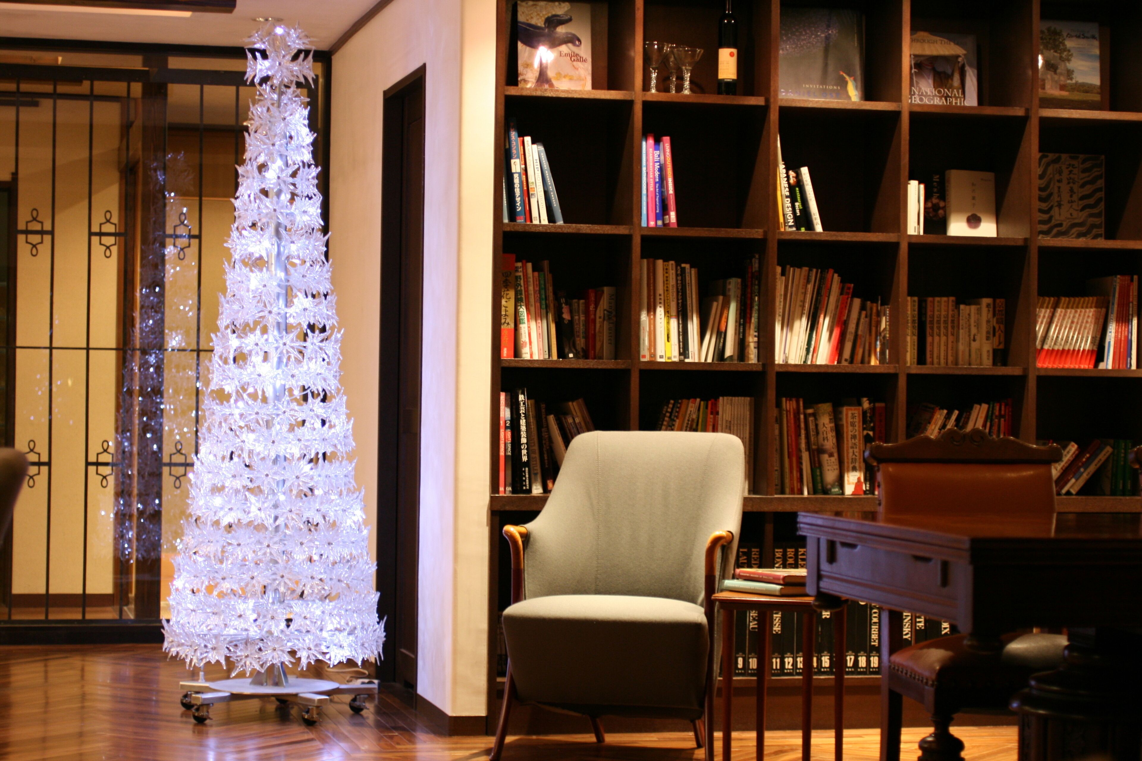 A room with a wooden bookshelf filled with books, a white Christmas tree, and a light-colored armchair.