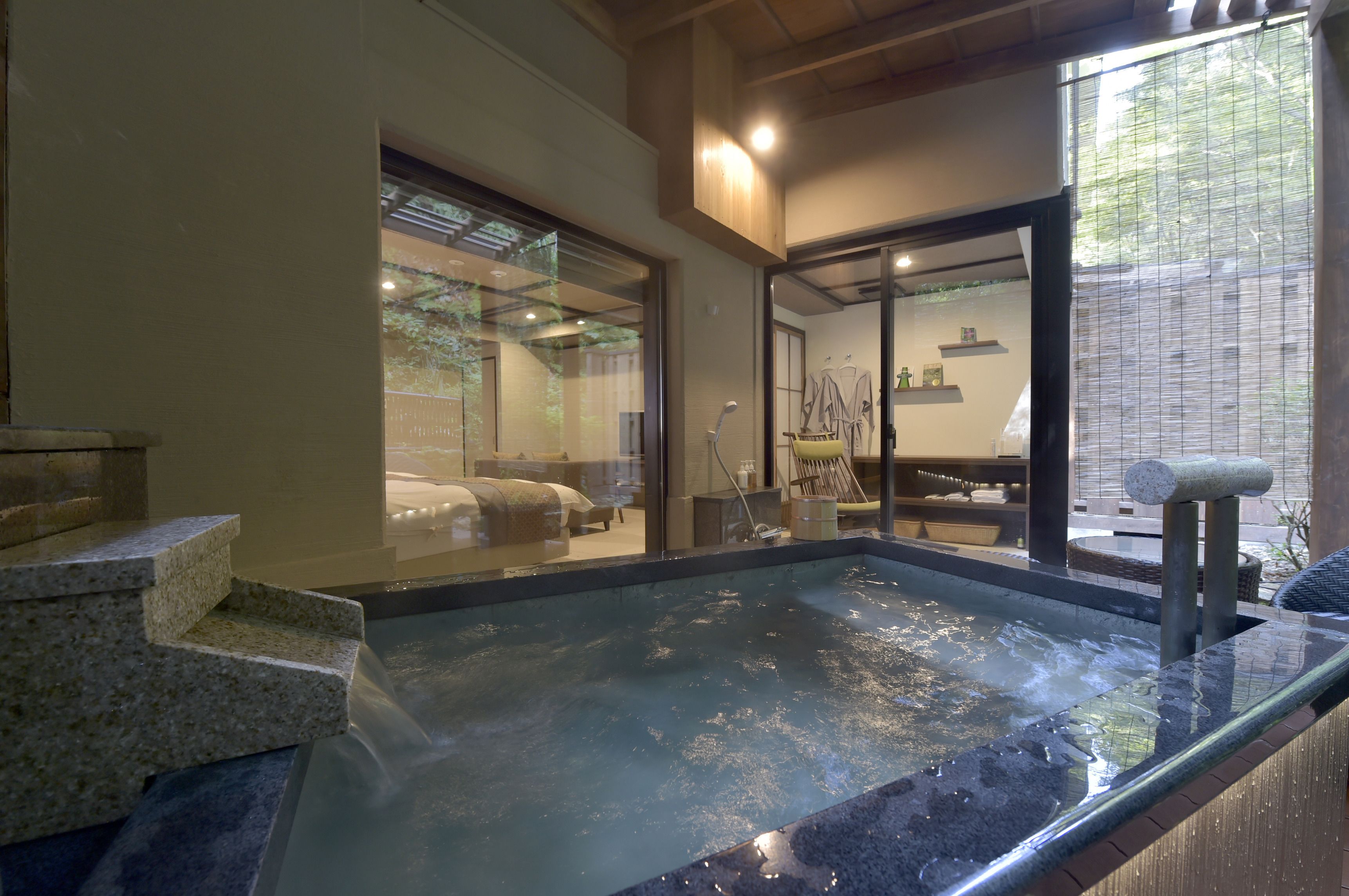 A hot tub area with steps leading into it, a glass door leading to a room with a bed, and a view of a garden outside.