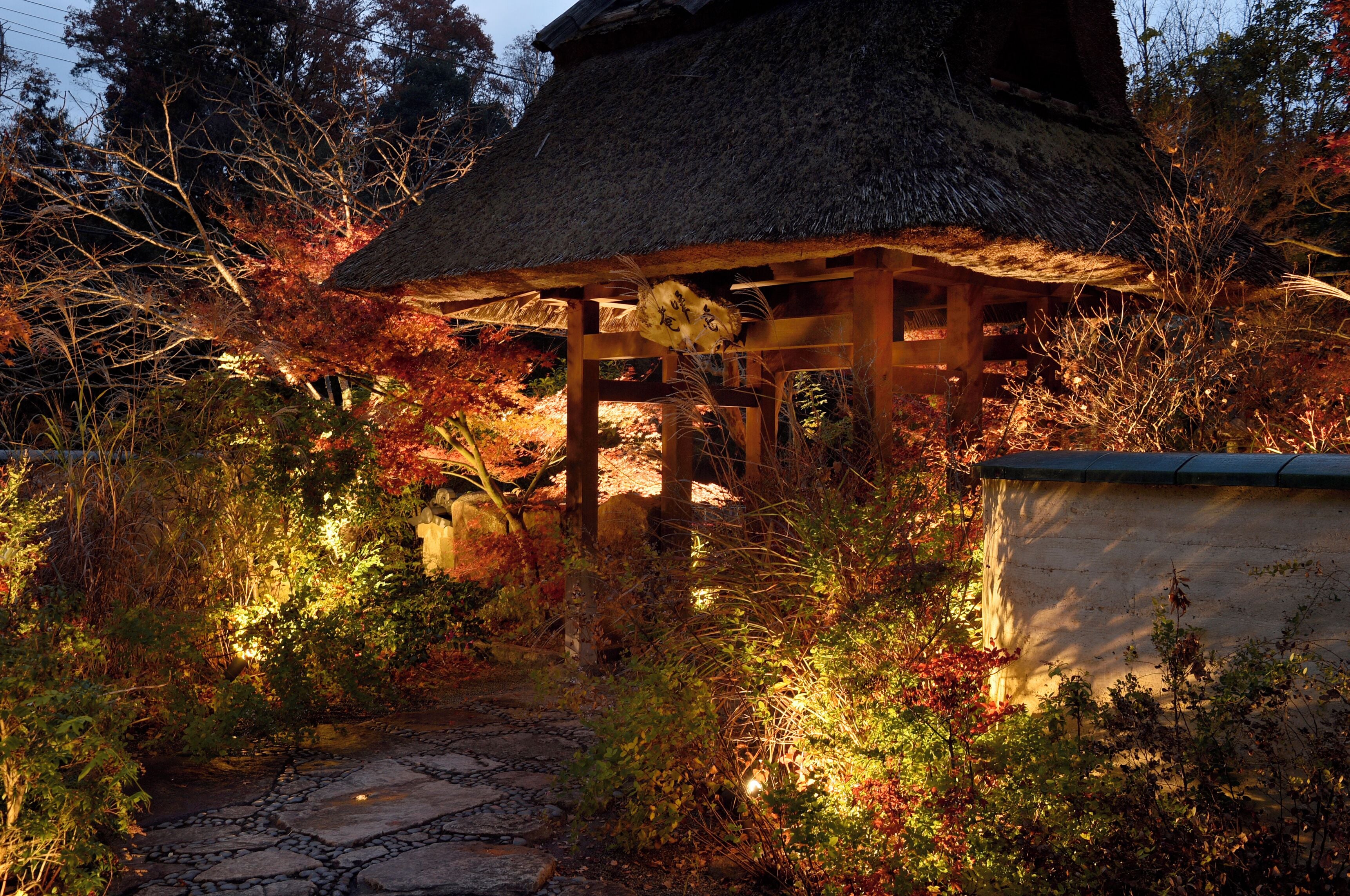 A traditional Japanese-style structure with a thatched roof, surrounded by autumn foliage and a stone pathway.