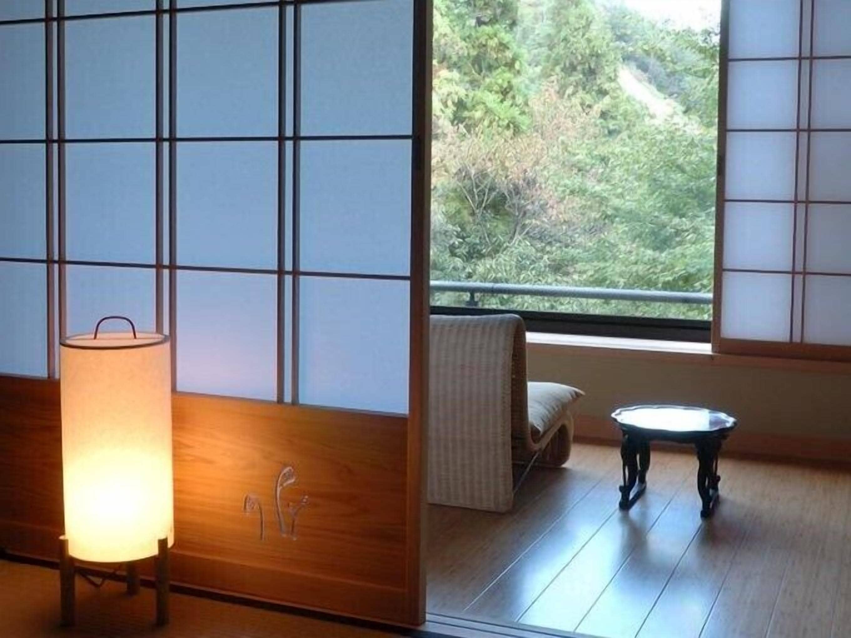Una habitación con esteras de tatami, suelo al estilo japonés tradicional, una mesa de madera, un cojín y vistas a la vegetación a través de una ventana.