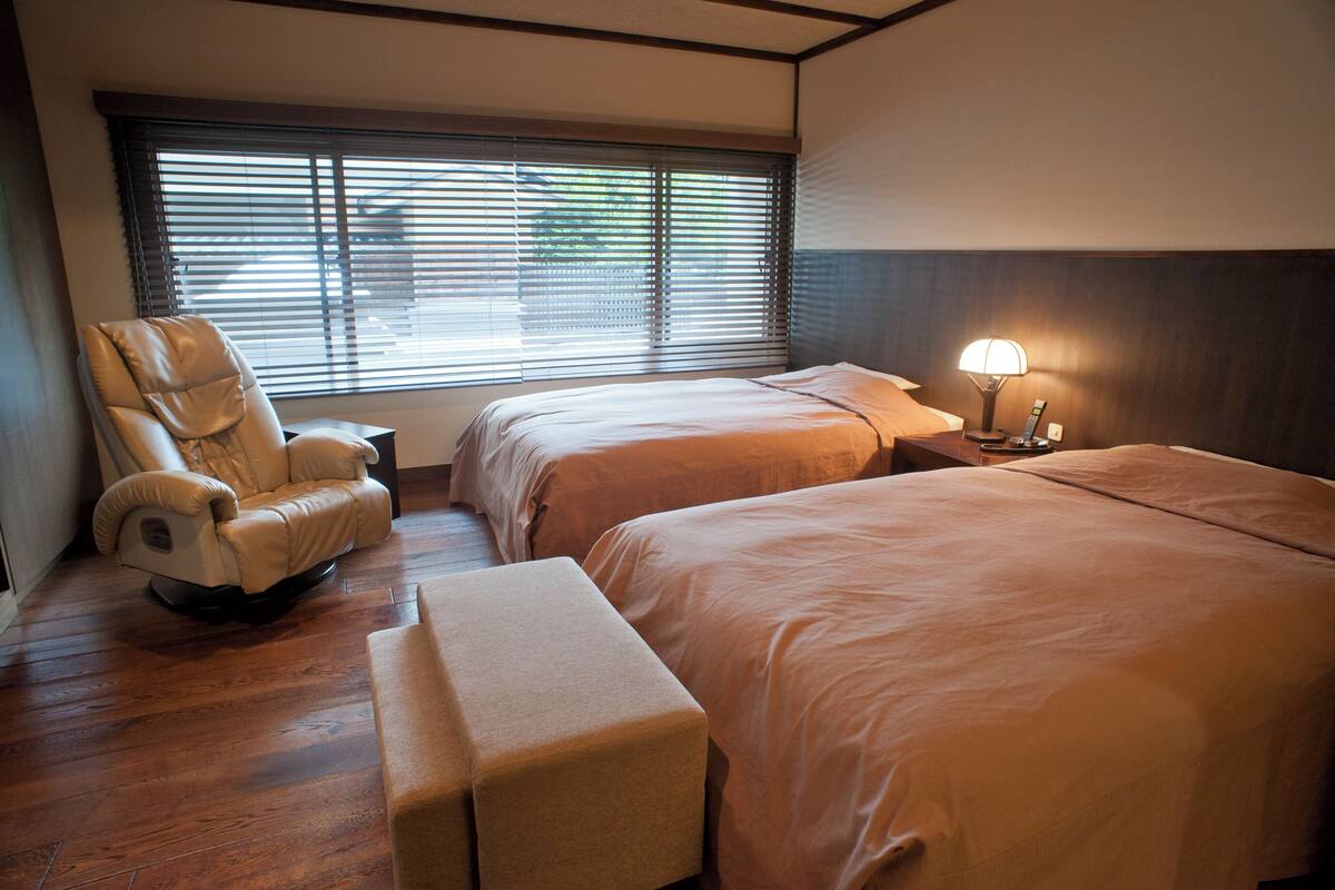 Habitación de hotel con dos camas, una silla de masajes, una mesita de noche con lámpara y una ventana con persianas.