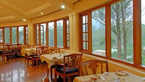 A dining area with wooden tables and chairs, large windows with a view of trees and hills. - KTDC Tea County Munnar (Devikolam)