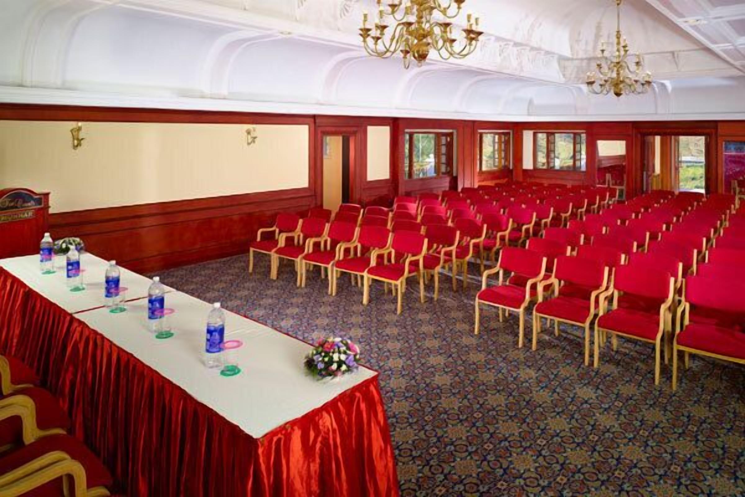 A conference room with rows of red chairs, a long table with water bottles and a flower arrangement, and a large screen at the front.