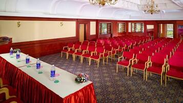 A conference room with rows of red chairs, a long table with water bottles and a flower arrangement, and a large screen at the front.