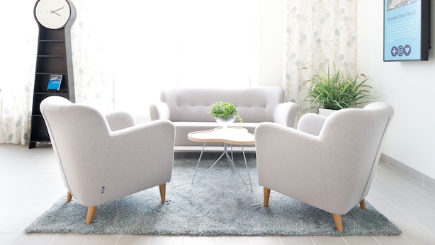 A modern living room with a sofa, two armchairs, a coffee table, a clock, a plant, and a framed informational poster. - Attendo Park Hotell (Huddinge)