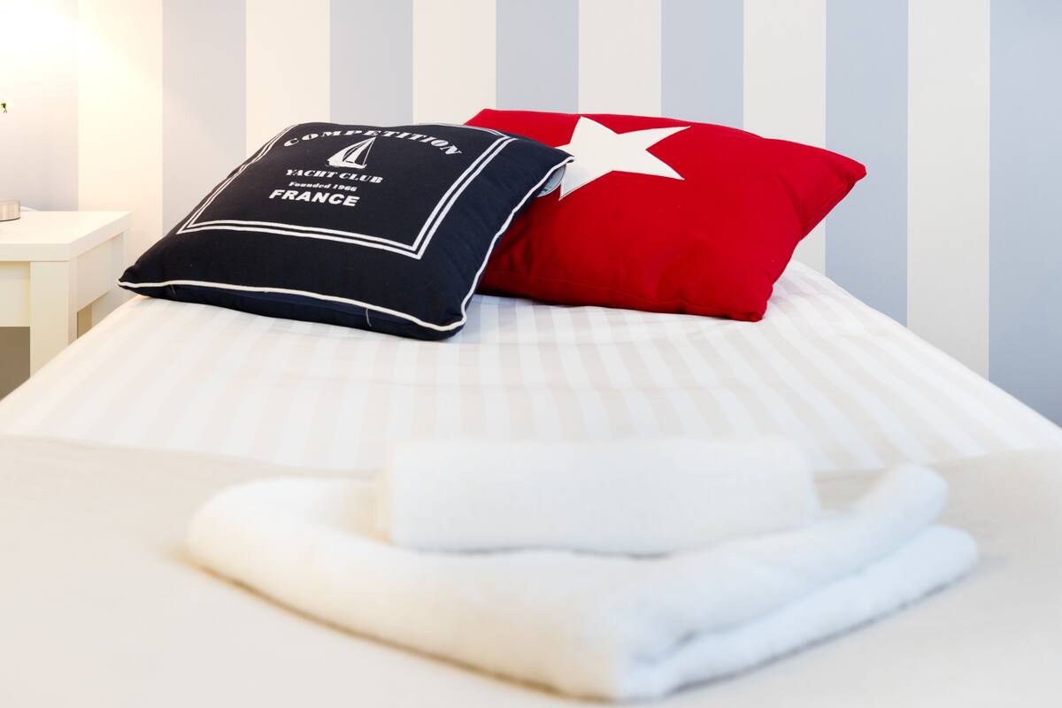 A neatly made bed with striped bedding, a red pillow, and a navy pillow with text.