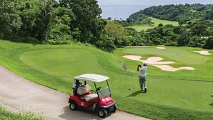 Un campo de golf con un carrito rojo, un golfista tomando un tiro y un caddie cercano.