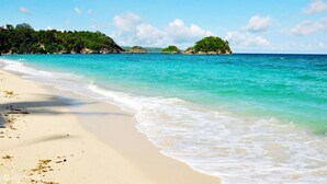 Una playa con agua turquesa cristalina, orilla de arena y colinas verdes al fondo.