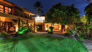 Un edificio de dos plantas con una entrada bien iluminada, un área de estar al aire libre techada y un jardín ajardinado con césped y árboles pequeños.